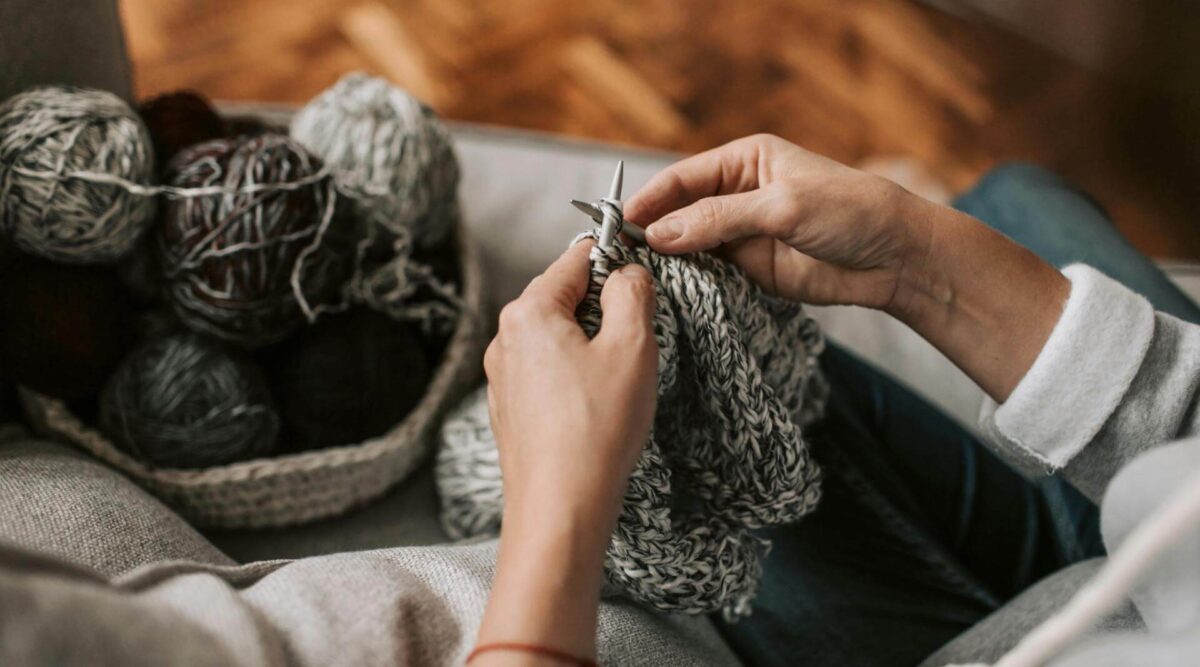a close up shot of a person knitting on circular needles. they are knitting with several strands held together and the resulting knit fabric is a grey melange colour. The shot is focused on the persons hand and their knitting project in the foreground and in the background there is a woven or crocheted basket holding the balls of yarn they are knitting from