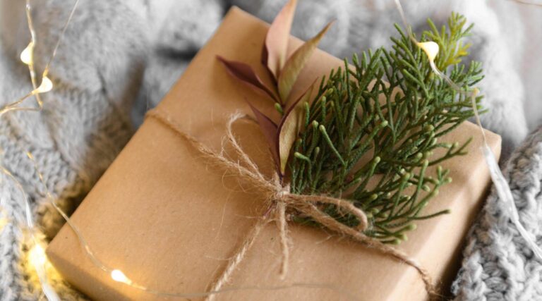a close up shot of a present wrapped in brown paper and string with a spring of christmas tree to decorate. The christmas gift is laid on a cable knit blanket and has fairy lights scattered around it